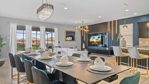 A modern dining area with a long table set for a meal, a television mounted on the wall, and a view of the outdoors through large windows.