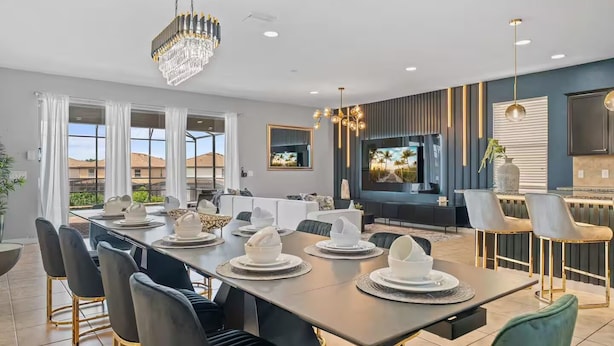 A modern dining area with a long table set for a meal, a television mounted on the wall, and a view of the outdoors through large windows. - FL- 1904 Nice Ct (Kissimmee)