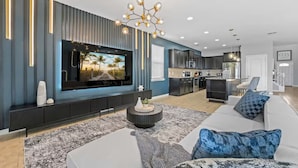 A modern living room with a flat-screen TV, a sofa, a coffee table, and a kitchen in the background. - FL- 1904 Nice Ct (Kissimmee)