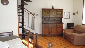 A living room with a wooden staircase, a dining table with chairs, a sofa, and a kitchen area with cabinets and a television. - Cozy Stay With Shared Pool (San Michele al Tagliamento)