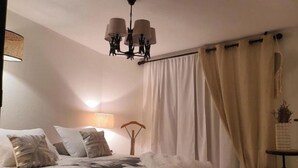 Un dormitorio con cama, lámpara colgante, mesita de noche con planta y ventana con cortinas.