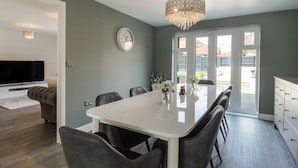 Un coin repas avec une table blanche, des chaises grises, un lustre et une horloge au mur.