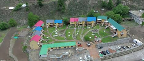 Vue aérienne d’un quartier résidentiel avec de nombreuses maisons, dont certaines ont des toits colorés, et un bâtiment central doté d’un toit végétalisé. On y voit des zones pavées, quelques voitures et un petit espace clos avec des sièges extérieurs.
