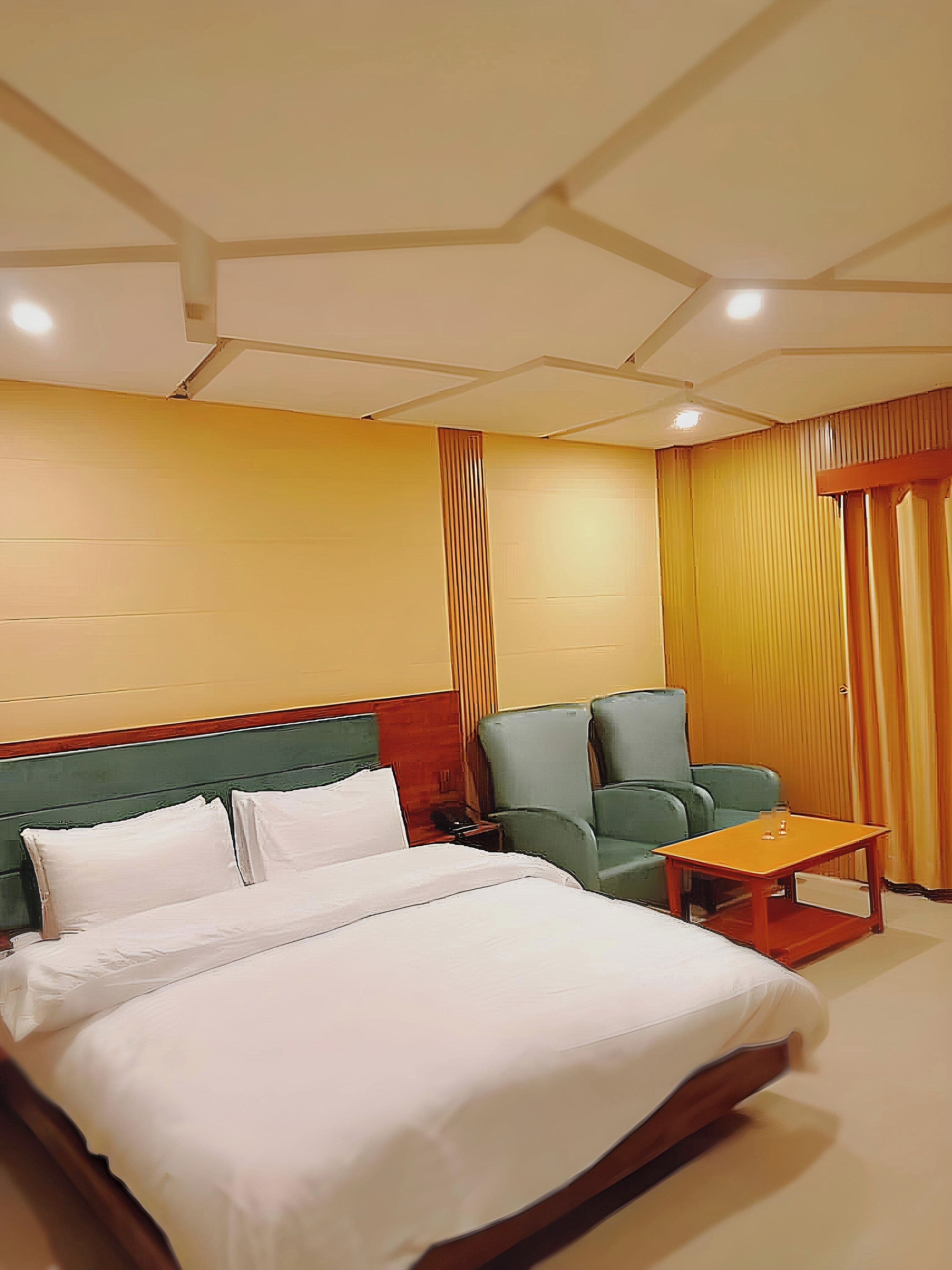 Una habitación de hotel con una cama grande, un sillón verde, una mesa de centro de madera y un cabecero de madera.