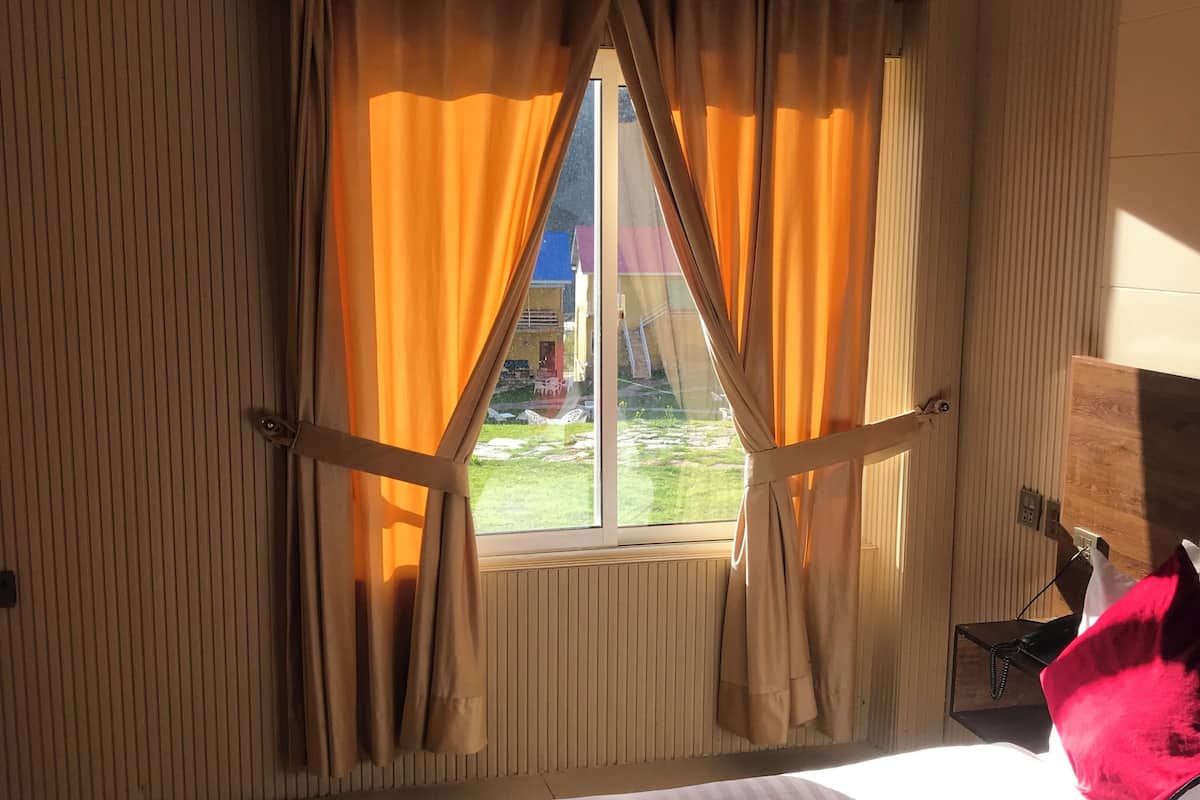 A hotel room with a large bed, a window with curtains, a radiator, and a small table with a lamp.