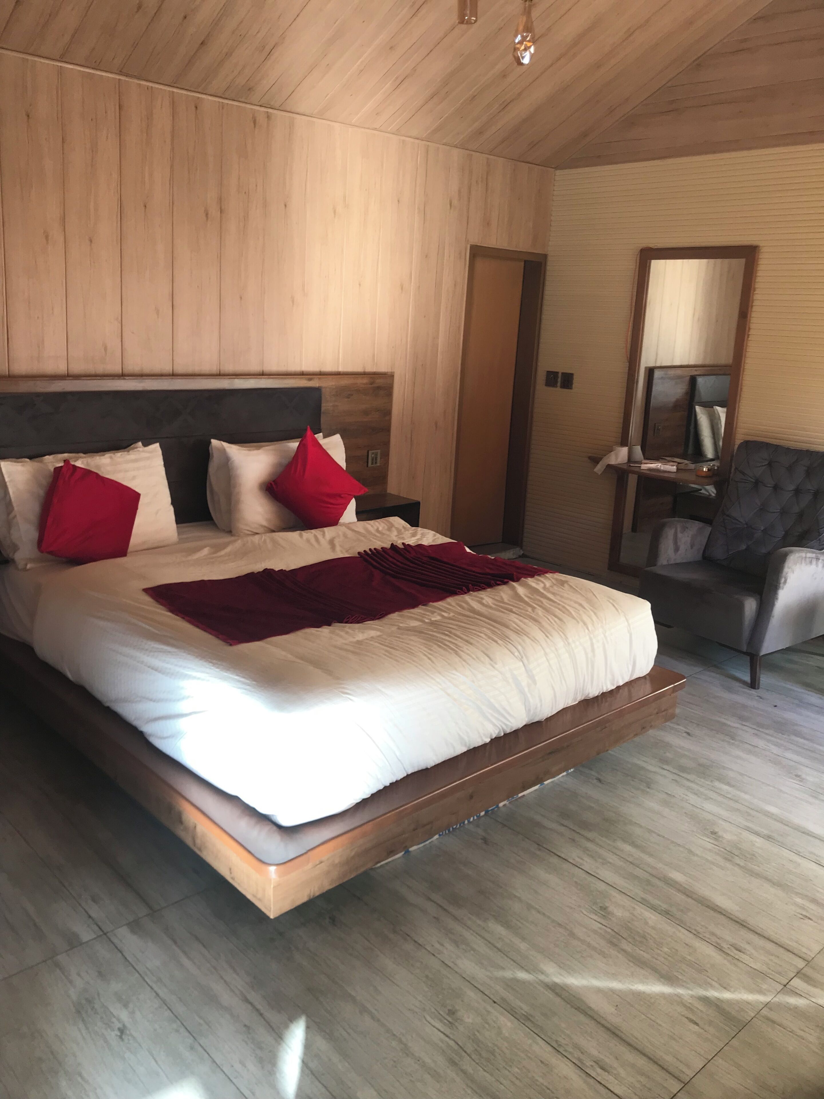 A modern bedroom with a wooden ceiling, a large bed with white and red bedding, a bedside table, and a chair.
