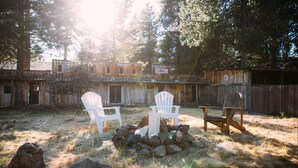 A rustic outdoor setting with a fire pit, two white Adirondack chairs, and a wooden chair.