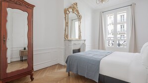 Une chambre avec une armoire en bois, un grand miroir décoratif, un lit recouvert d’une courtepointe bleue et une vue sur un balcon par la fenêtre.