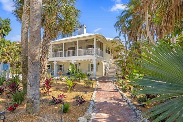 Uma casa de dois andares com varanda coberta, rodeada por um jardim com diversas plantas e um caminho de tijolos.