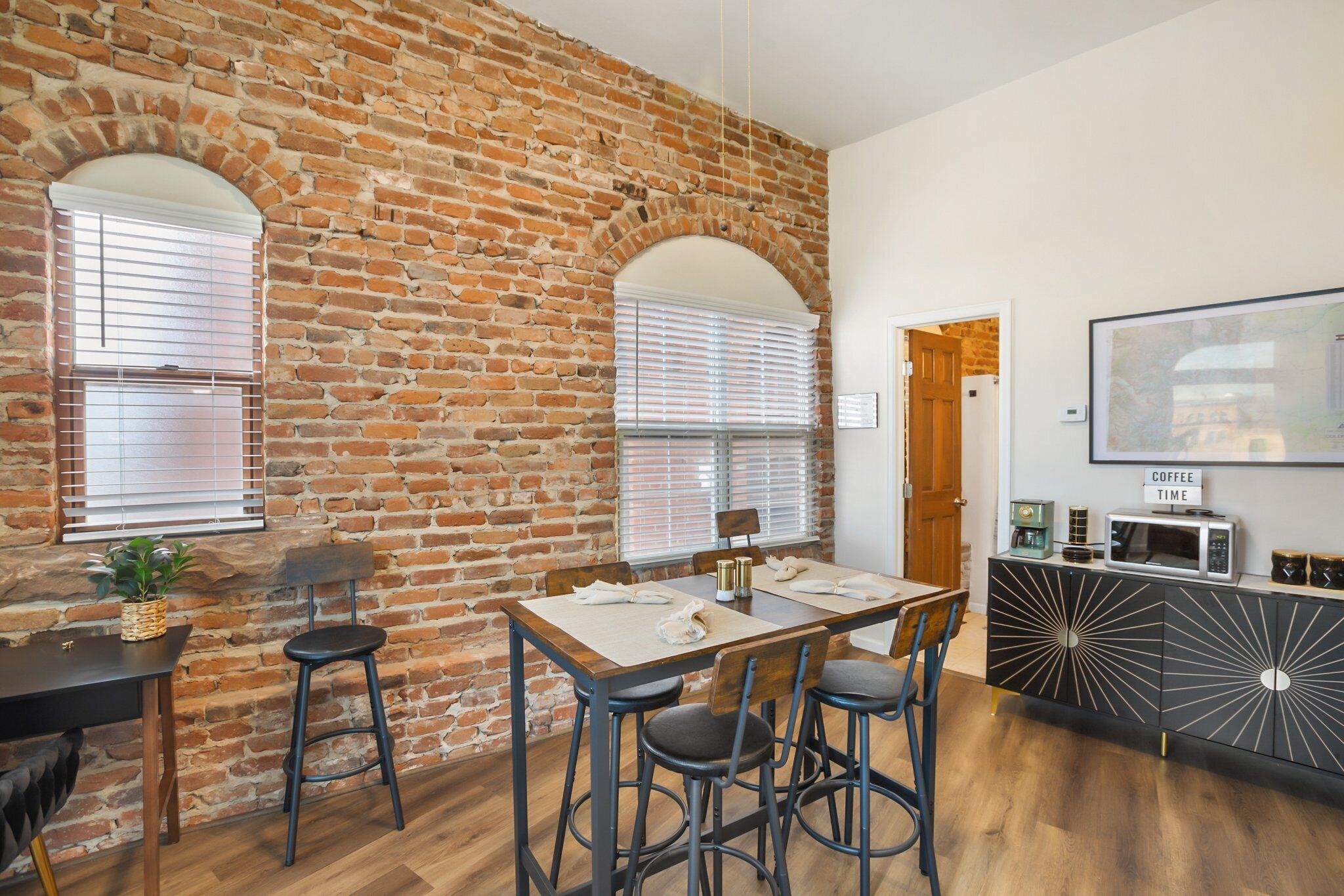 Un coin repas avec un mur en briques, une table en bois et des chaises. On y trouve une porte qui mène à une autre pièce et un meuble noir orné d’un motif décoratif.