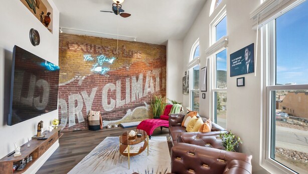 A modern living room with a large brick wall mural, a leather sofa, a flat-screen TV, and a large window offering a view of the outside. - The Ghost Lounge (Victor)