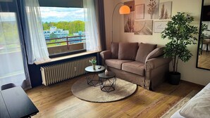 A modern room with a brown sofa, a small round table with a vase, a potted plant, and a view of buildings through a window with sheer curtains.