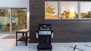 Ein moderner Grillbereich im Freien mit einer Bank, einem Glastür-Eingang und Blick auf ein Gebäude mit großen Fenstern.