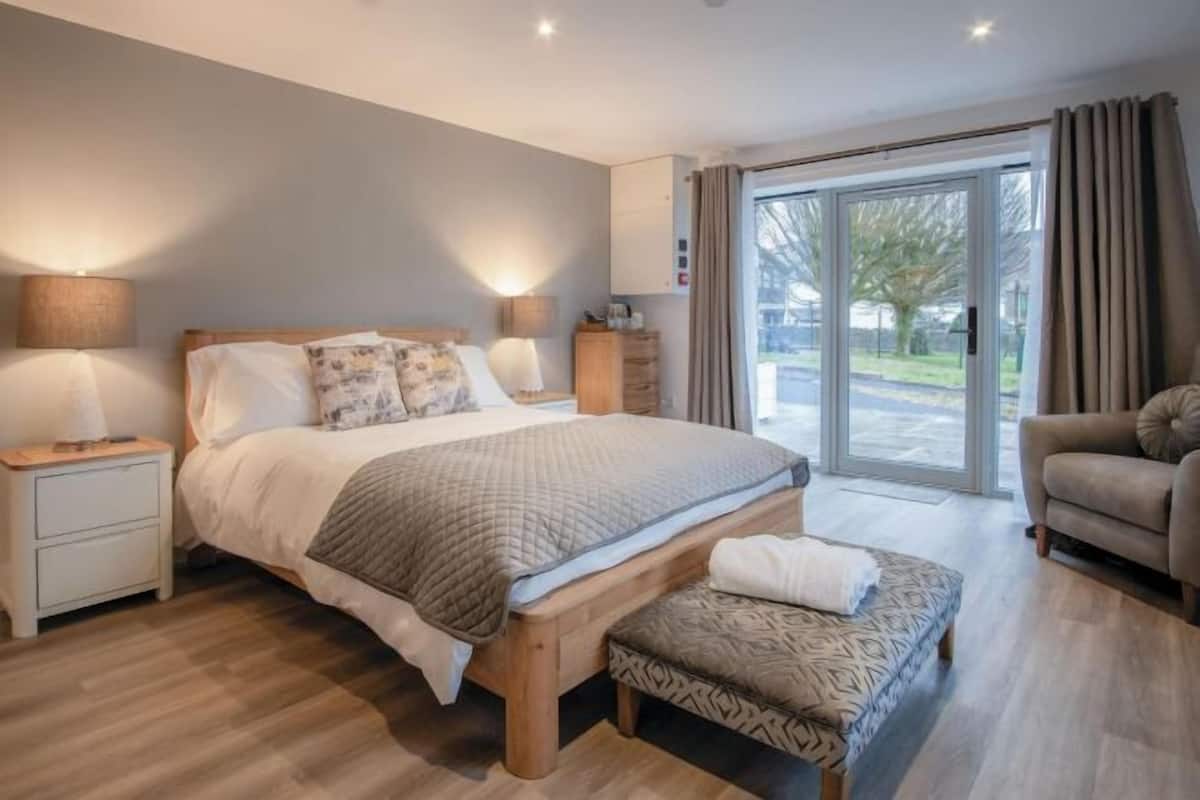 Een moderne slaapkamer met een groot bed, nachtlampjes, een bankje en uitzicht op het buitenlandschap door een glazen deur.