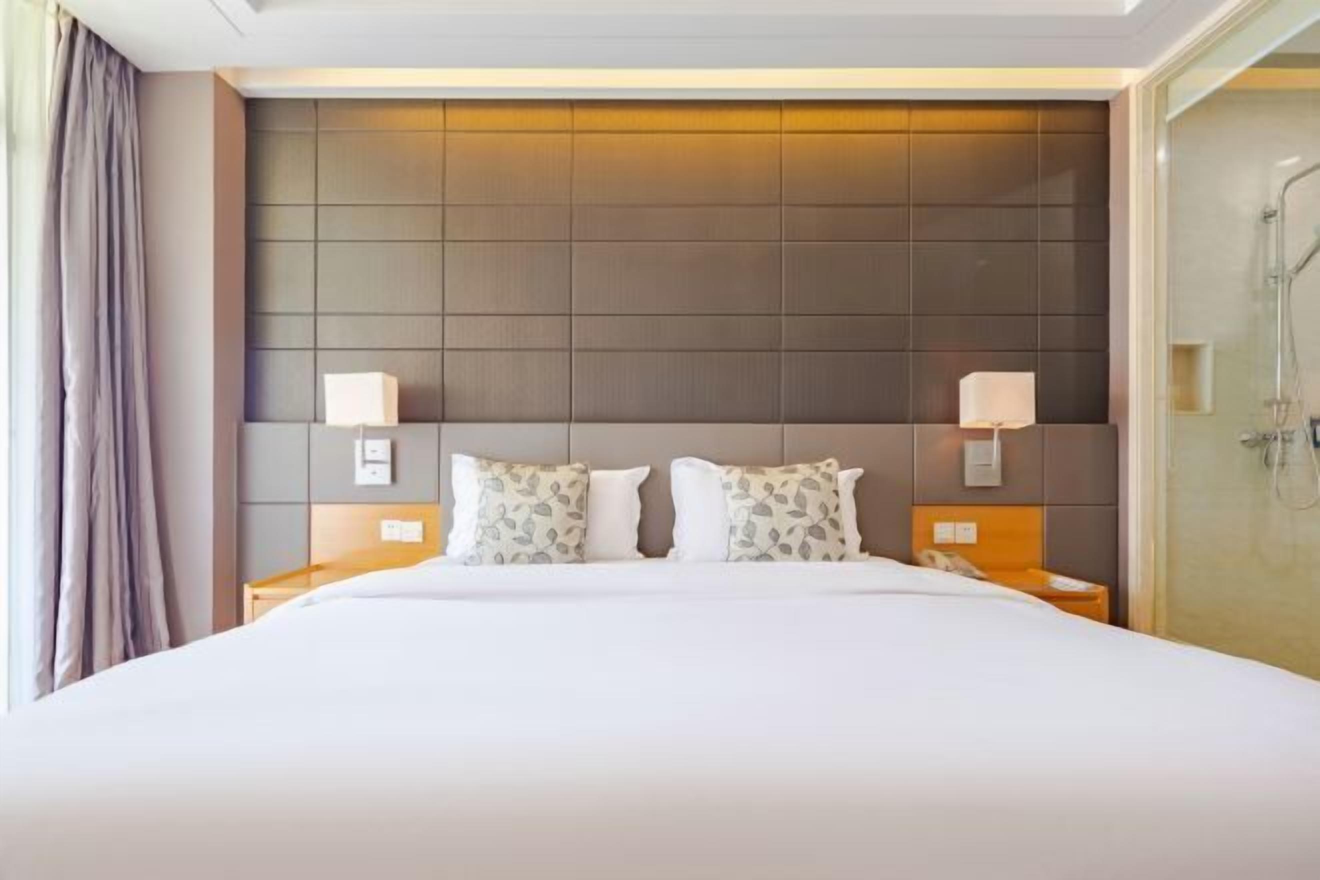 Hotelkamer met een groot bed, twee nachtkastjes met lampen, een hoofdbord met paneeldecoratie en een douchecel.
