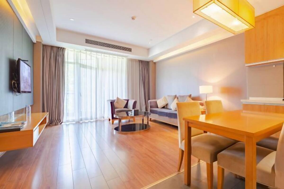 Una habitación de hotel moderna con piso de madera, un conjunto de sofás, una mesa de comedor y un televisor montado en la pared.
