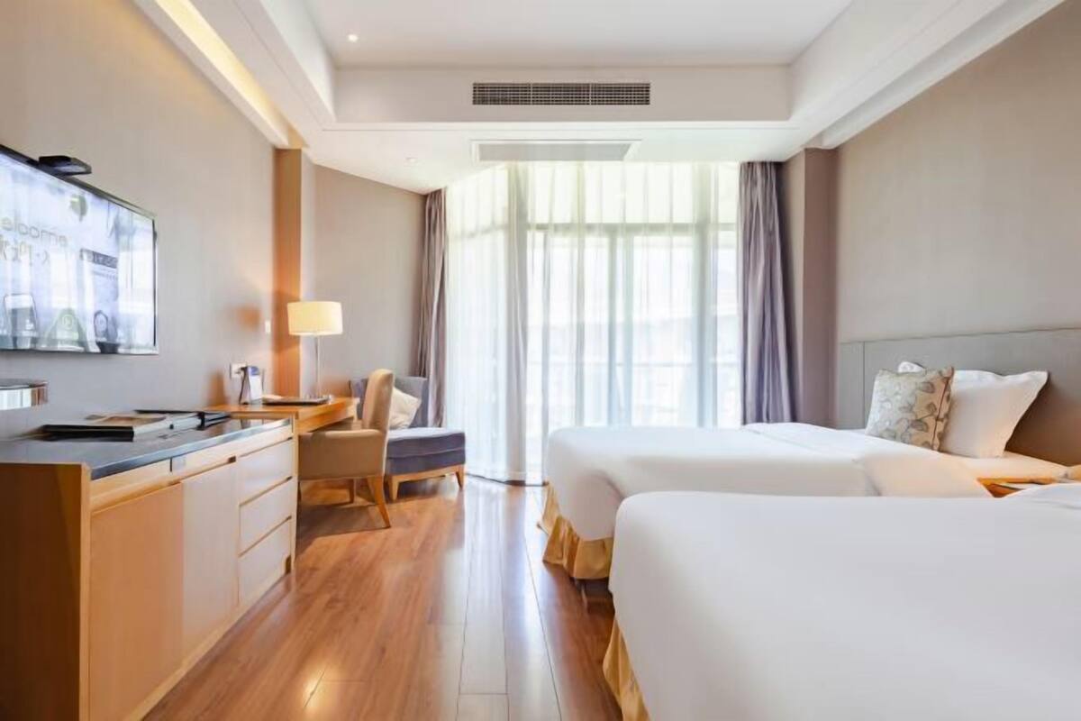 Habitación de hotel con cama, escritorio, silla, televisor y un amplio ventanal con cortinas.