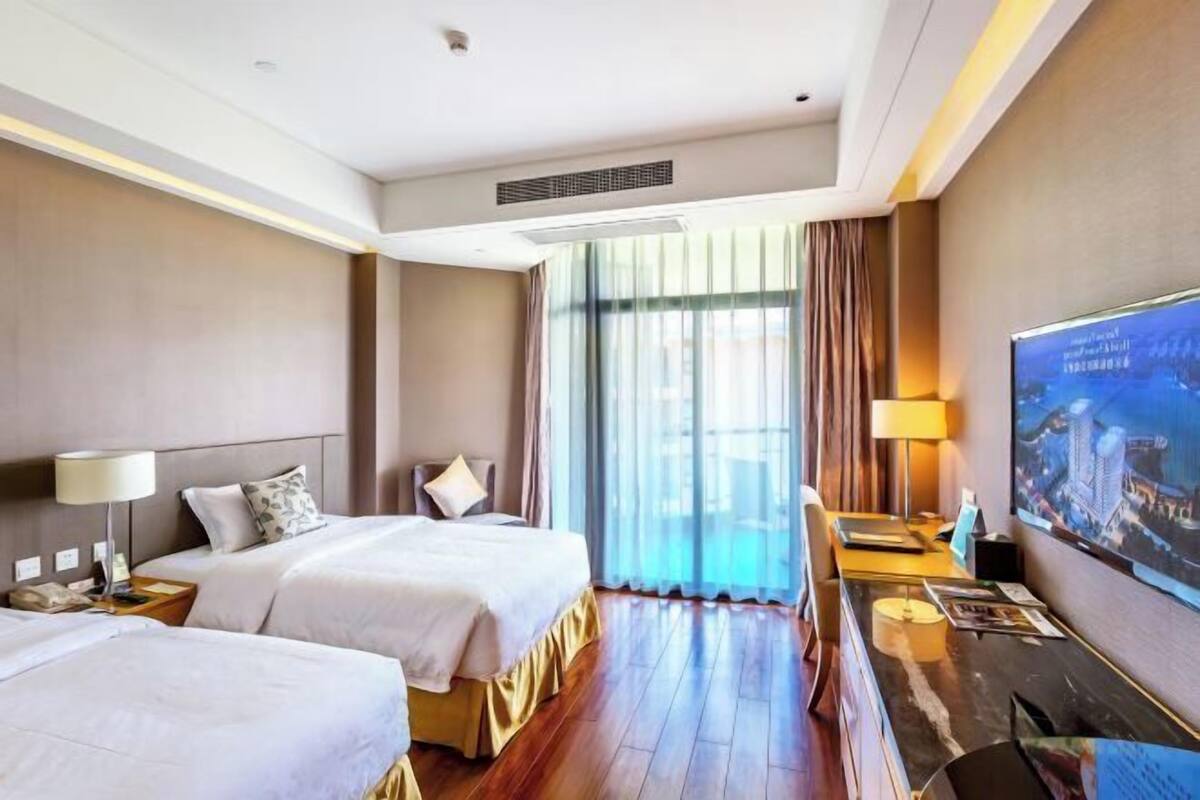 Habitación de hotel con dos camas, un escritorio, un televisor y un ventanal con cortinas.