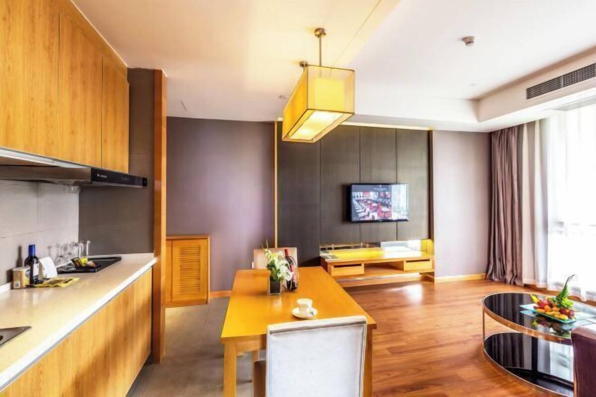 Una habitación de hotel moderna con una mesa de comedor de madera, un sofá, un televisor montado en la pared y un amplio ventanal con cortinas.