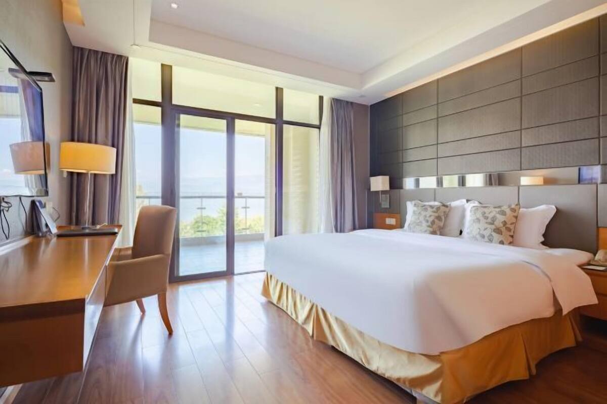 Una habitación de hotel moderna con una cama grande, un escritorio y una silla. Cuenta con un balcón con cortinas y vistas al exterior.