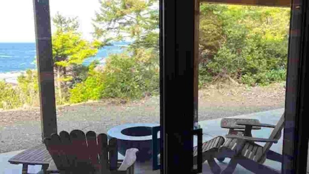 A covered outdoor seating area with a table and chairs, overlooking a scenic view of the ocean and greenery. - The Suite at Sunset Point (Ucluelet)