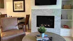 A living room with a fireplace, a flat-screen TV mounted above it, a bookshelf, and a coffee table with books and a plant. - The Suite at Sunset Point (Ucluelet)