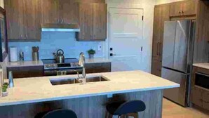 A modern kitchen with a large island, bar stools, stainless steel appliances, and wooden cabinets. - The Suite at Sunset Point (Ucluelet)