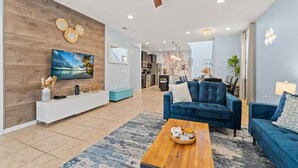 A modern living room with a TV, blue sofas, a wooden coffee table, and a kitchen in the background.