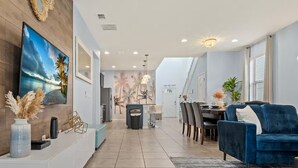 A modern living room with a blue sofa, a TV on the wall, and a dining area with a table and chairs.