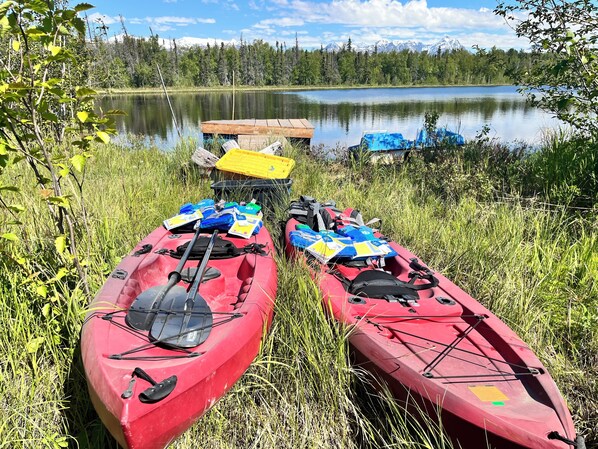Two red kayaks with paddles and life jackets on a grassy shore by a calm lake with a dock in the background.