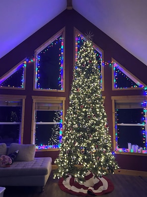 A Christmas tree decorated with lights and ornaments, positioned in a room with a sloped ceiling and large windows. - Mountain-View Retreat: Pond, Kayaks, Fire Pit, Hot Tub – Palmer, Alaska (Palmer)