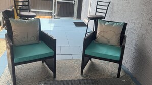Une terrasse avec deux fauteuils en osier dotés de coussins turquoise, une table noire ornée d’une figurine de tortue, et une vue sur une clôture en bois et des arbres.