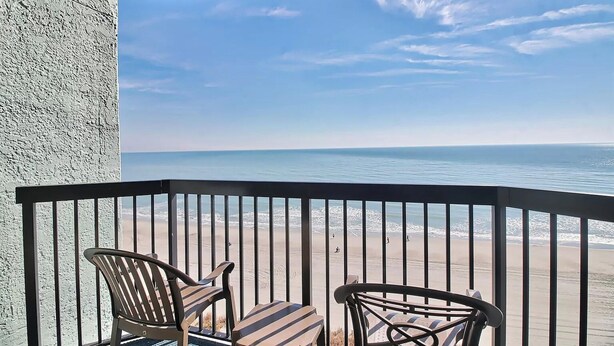 A balcony with two chairs and a table, overlooking a beach with waves and a clear sky.