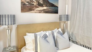 Une chambre à coucher comprenant un lit, deux lampes de chevet, une tête de lit et un tableau représentant un paysage côtier, accroché au mur.