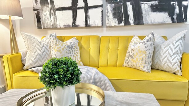 Un canapé jaune orné de coussins à motifs, un vase blanc contenant des feuilles vertes et un plateau doré.