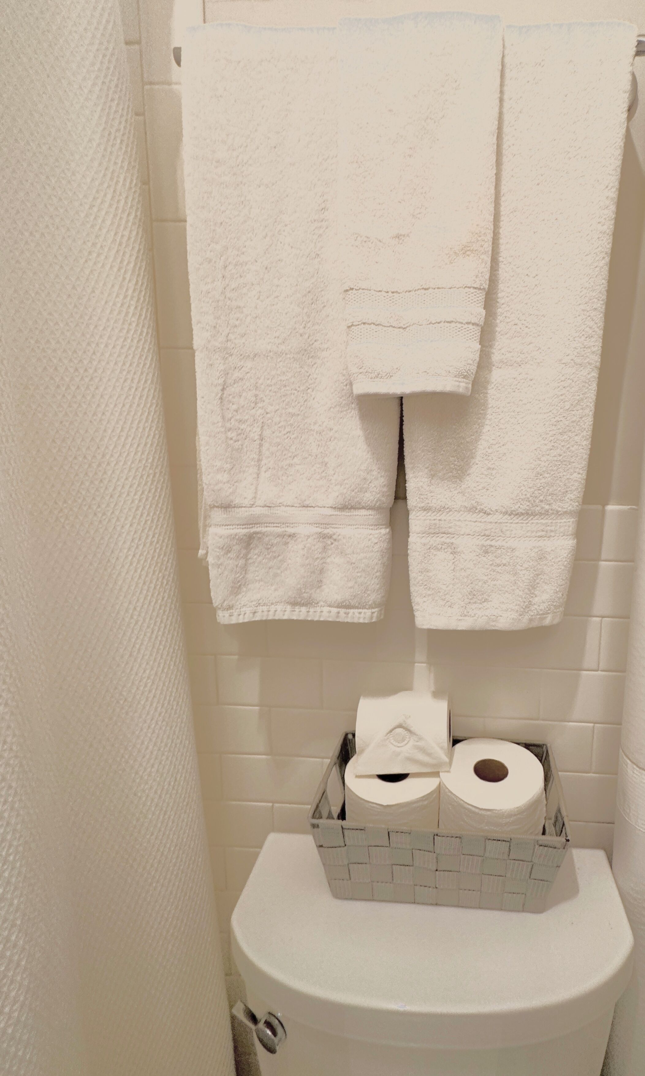 A bathroom with a white toilet, a white towel hanging on the wall, and a basket containing toilet paper and a roll of paper towels.