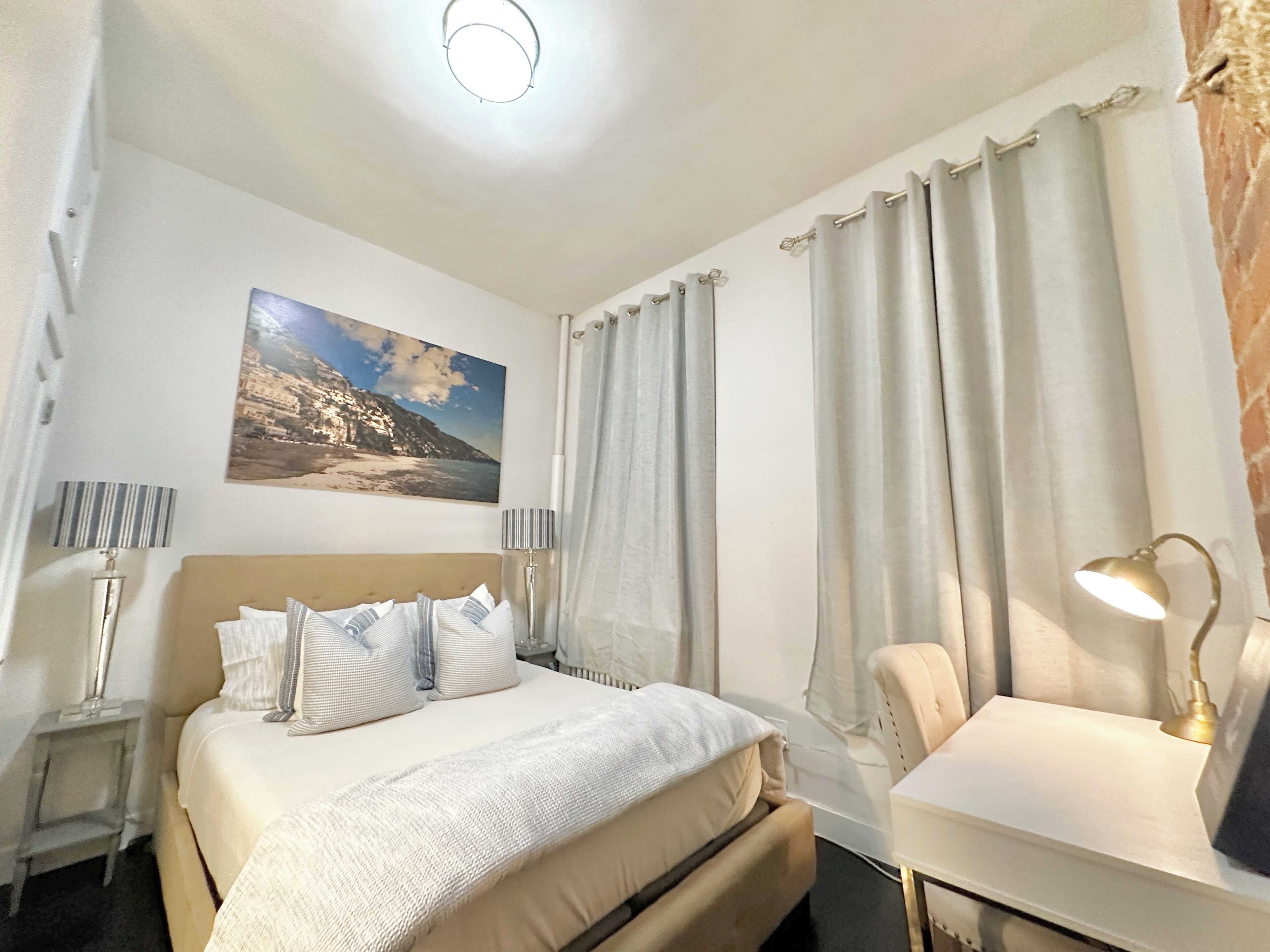 Une chambre d’hôtel avec un lit, un bureau, une lampe, des rideaux et un tableau au mur.