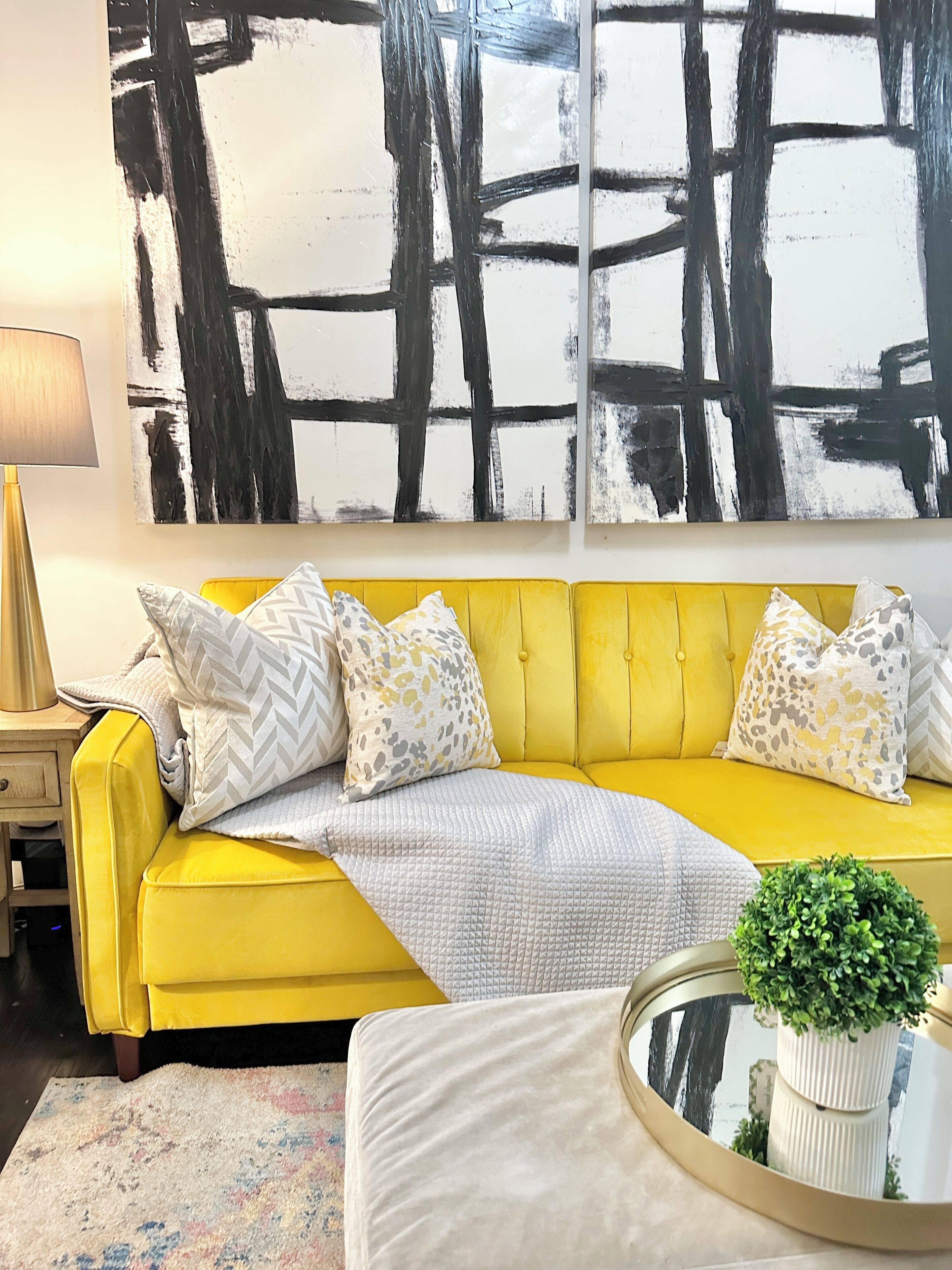 A yellow sofa with patterned pillows, a small round table with a plant, and a large abstract painting on the wall.