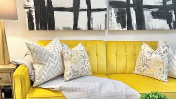 A yellow sofa with patterned pillows, a small round table with a plant, and a large abstract painting on the wall.