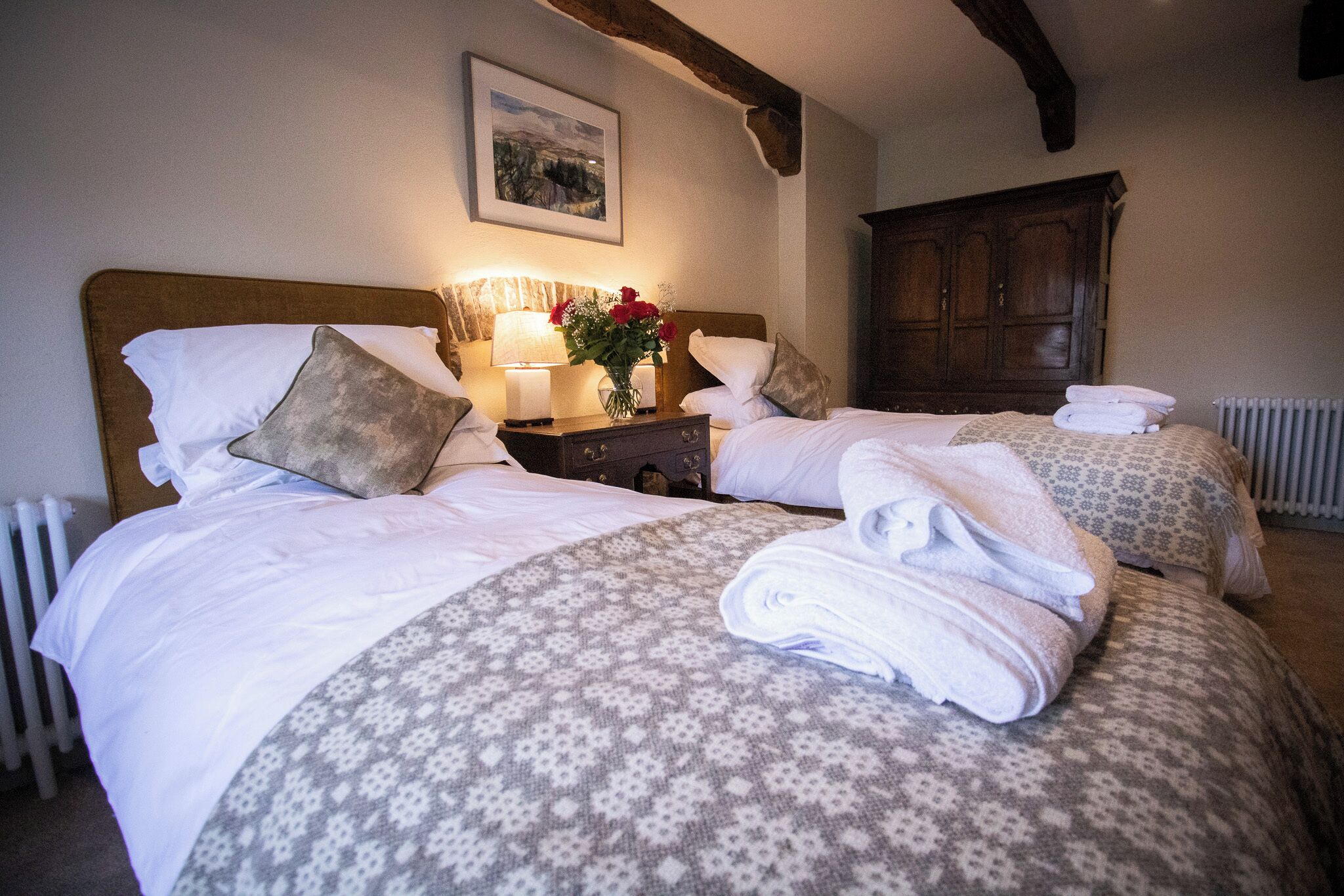 Een hotelkamer met twee bedden, elk met een beddekleed met dessin, kussens en opgevouwen handdoeken. Er staat een nachtkastje met een lamp en een bloemenvaas. Een houten kledingkast is op de achtergrond te zien.