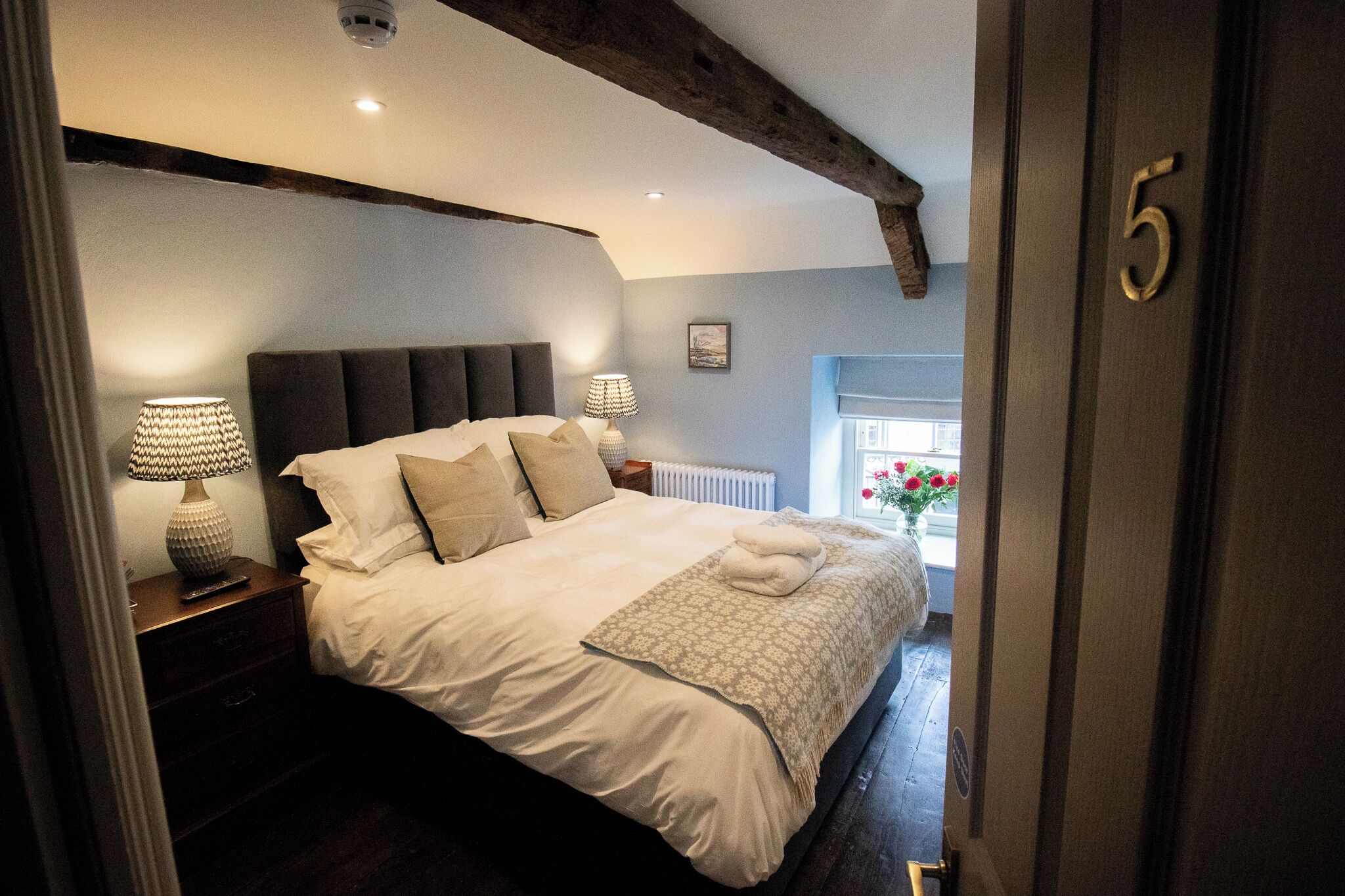 Un dormitorio con una cama grande, dos lámparas de noche, una mesita de noche, una ventana con flores y una puerta con el número 5.