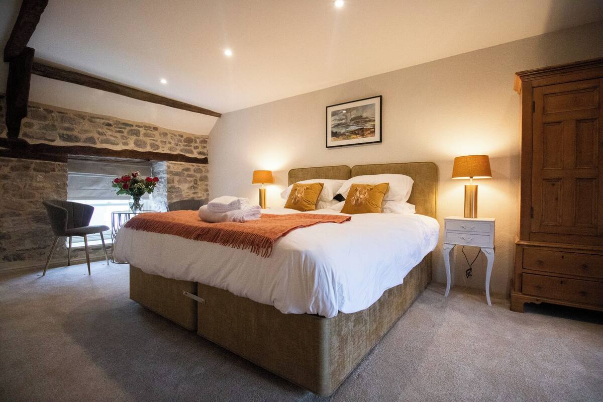 Un dormitorio con una cama grande, dos mesitas de noche con lámparas, un armario de madera, una pared de piedra y una silla.