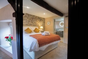 A bedroom with a stone wall, a bed with white bedding and a red throw, a nightstand with a lamp, and a view of the bathroom through an open door. - Y Castell  (Llangadog)