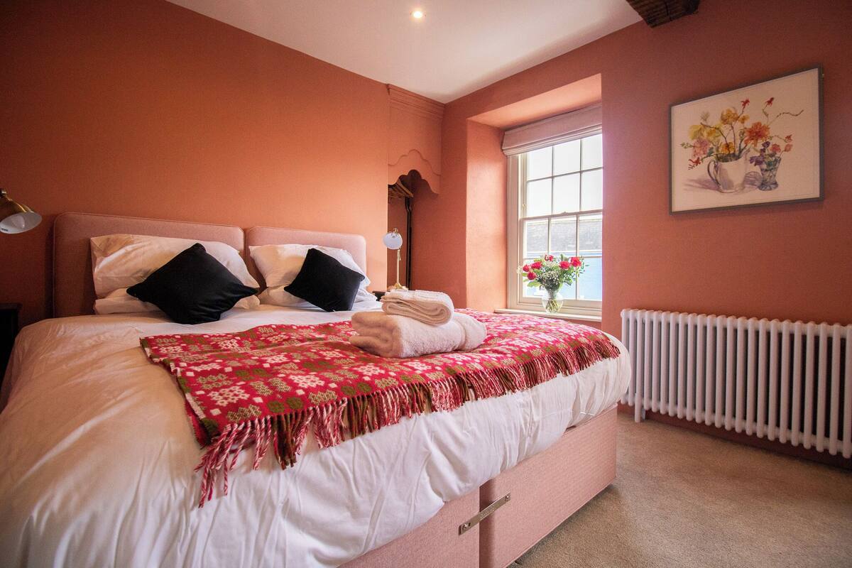 Un dormitorio con una cama, almohadas, una lámpara de noche, una ventana con flores y un radiador.