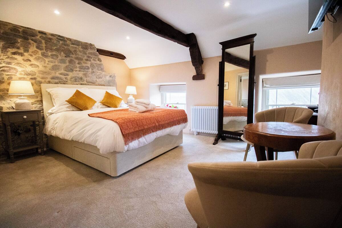 Un dormitorio con pared de piedra, una cama con sábanas blancas y almohadas naranjas, una mesita de noche con lámpara, una mesa redonda de madera y una silla.