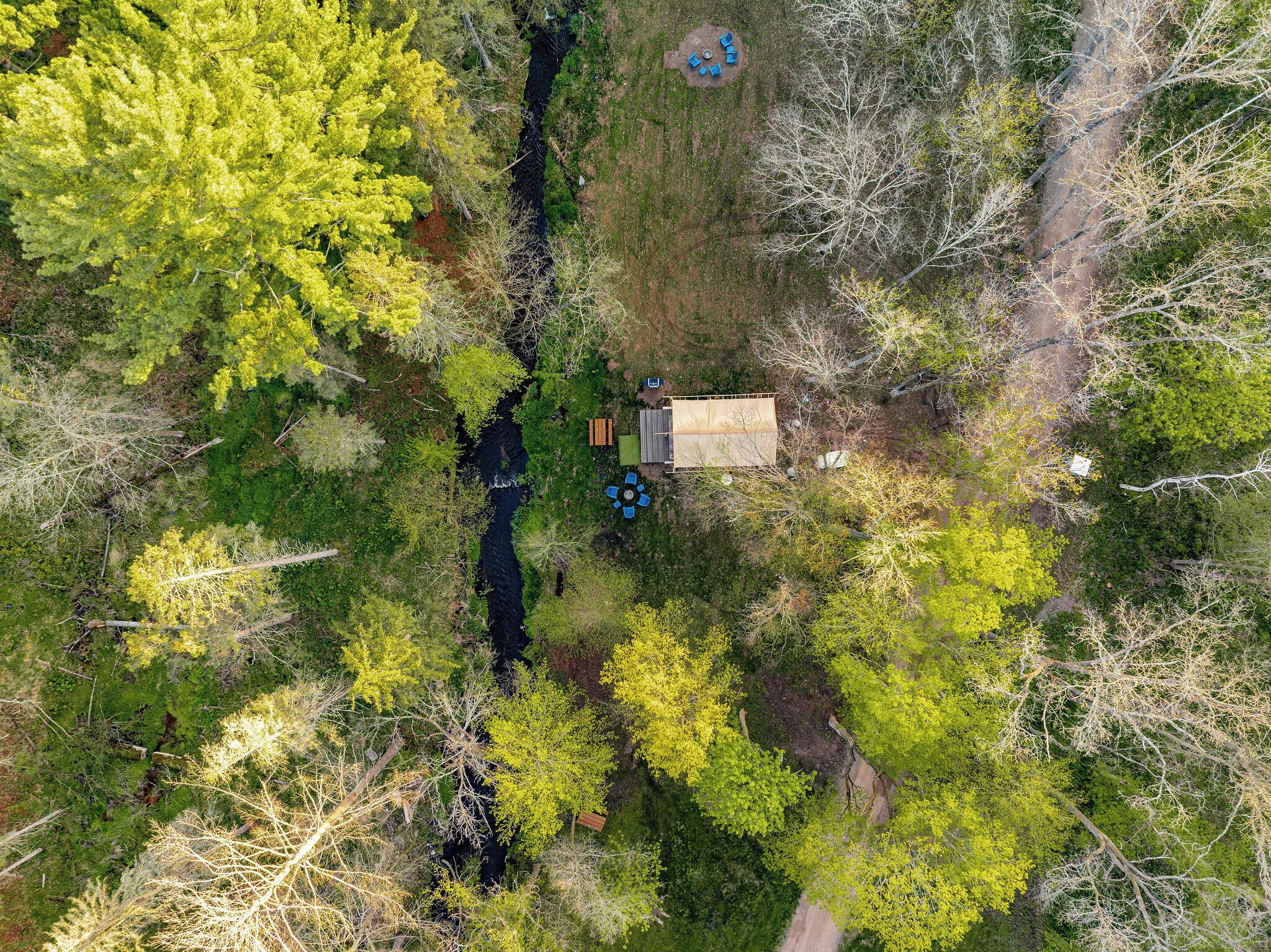An aerial view of a cabin surrounded by dense forest, with a small stream running through the property.