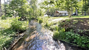 Una baita con un portico e un piccolo balcone, immersa in una fitta foresta e affiancata da un ruscello.