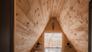 Ein gemütliches Schlafzimmer im Dachgeschoss mit schrägen Wänden, einem Bett mit weißen Leinen, einem flauschigen Teppich und einem Fenster mit Blick auf Bäume.
