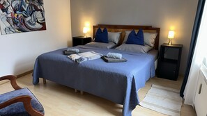 Un letto rifatto con una coperta blu, due cuscini e due asciugamani piegati. Sono presenti due comodini con lampade, una sedia di legno e un tappeto sul pavimento.