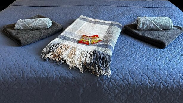 A double bed with a blue quilt, two pillows, two nightstands with lamps, and a folded towel on each side.